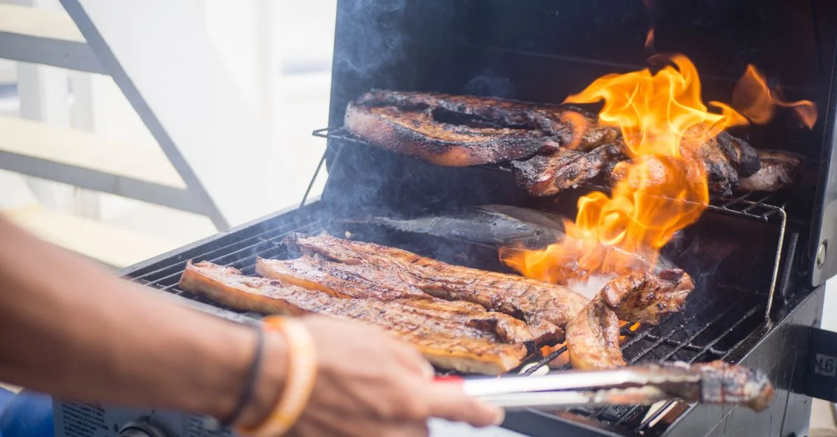 Une personne fait griller de la viande sur un barbecue à l'extérieur, les flammes étant visibles.
