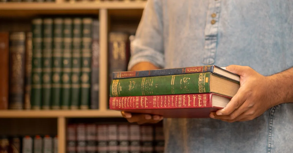 Dans une bibliothèque, un homme vêtu d'une chemise en jean tient des livres arabes empilés, symbolisant l'étude et l'apprentissage.