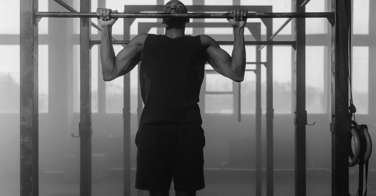 Photo en noir et blanc d'un homme réalisant des tractions dans une salle de gym intérieure, mettant en avant la force et la condition physique.